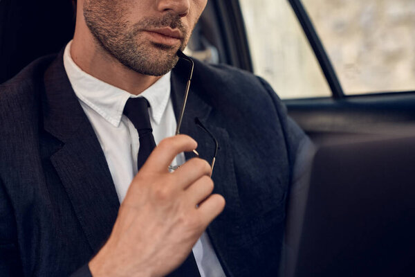 Cropped shot of thoughtful young man in full suit working on his laptop and holding his eyewear in hand while sitting in the car.