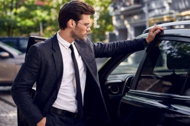 Formalite ve göz giysisi içinde kendine güvenen bir iş adamının arabaya yaslanıp başka yere bakması..