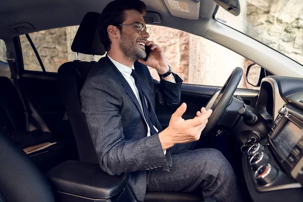 Nice business talk. Handsome young businessman talking on his smart phone and smiling while sitting on the front seat.