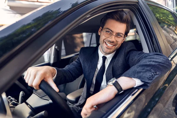 Happy young man in full suit and eyewear smiling while driving a car.