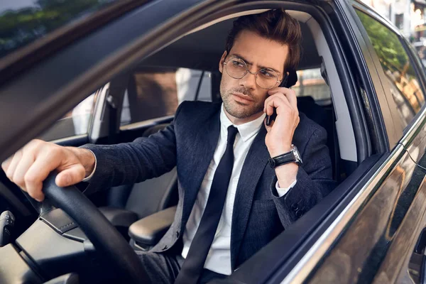 Nice business talk. Handsome young businessman talking on his smart phone and smiling while sitting on the front seat.