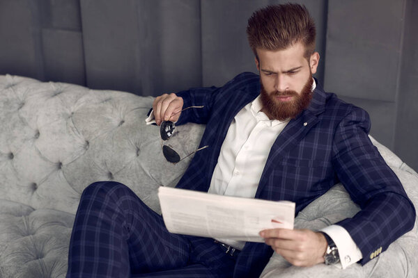Handsome stylish man in blue suit in a cage at home sitting on sofa and reading fresh newspaper
