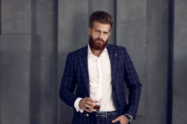 Takım elbiseli yakışıklı adam elinde bir bardak viskiyle kameraya bakıyor.