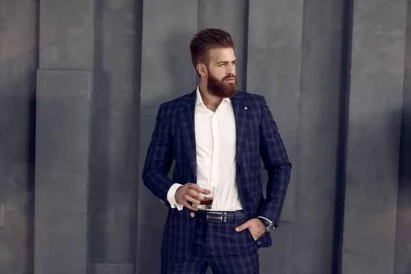 Handsome stylish man in suit stands with glass of whiskey, looking away