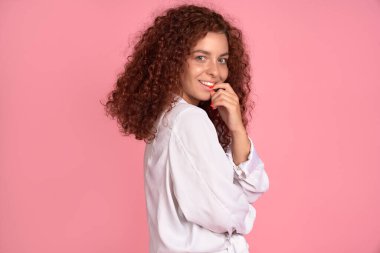 Uçan kıvırcık saçlı güzel kızıl saçlı kız pembe arka planda kameraya bakıp gülümsüyor.