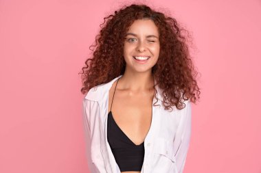 Uçan kıvırcık saçlı güzel kızıl saçlı kız pembe arka planda kameraya bakıp gülümsüyor.