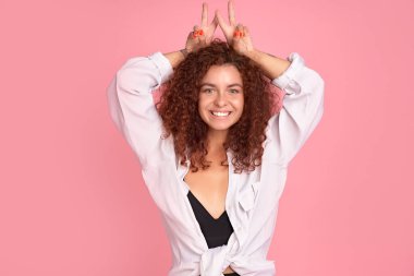 Yaşam tarzı ve güzel insanlar konsepti. Kapalı mekanda, kızıl saçlı, cilveli ve parmaklarını tavşan kulağı ya da boynuzları gibi başının arkasında tutan, flört ederken dilini gösteren şık bir kadın resmi..