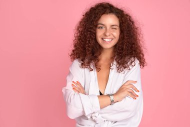 Uçan kıvırcık saçlı güzel kızıl saçlı kız pembe arka planda kameraya bakıp gülümsüyor.