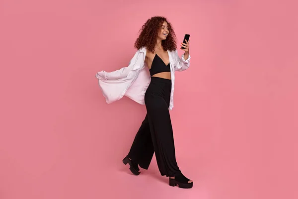 Full length profile photo of positive redhead girl use smartphone, type sms on social network wear style outfit pants high-heels isolated on pink color background...