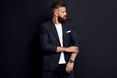 Mükemmel saç ve sakallı yakışıklı adam siyah arka planda dikilirken kollarını düzeltiyor..