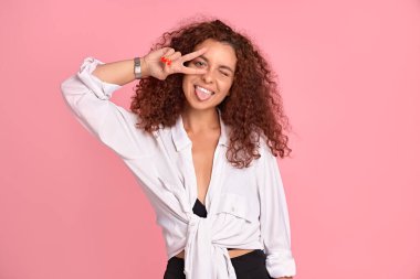 Gülümseyen bayan öğrenci, pembe duvara karşı poz verirken mutlu bir ifade takınıyor. V işaretini eliyle gösteren pozitif bir kız, boş zaman geçirmekten zevk alıyor. Yaşam tarzı, güzellik ve gençlik.