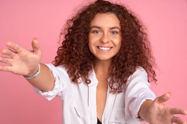 Bana sımsıkı sarıl. Kaygısız ve arkadaş canlısı kızıl saçlı hoş bir portre, pembe arka planda gülümseyen bir arkadaşa sarılmak için kameraya doğru elini çekiyor..