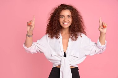 Dost canlısı görünüşlü, kendine güvenen, beyaz saçlı, sıradan giyinmiş, işaret parmağı kaldıran, yön gösteren, pembe arka planda reklamını yapan kızıl saçlı kız..