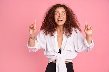 Dost canlısı görünüşlü, kendine güvenen, beyaz saçlı, sıradan giyinmiş, işaret parmağı kaldıran, yön gösteren, pembe arka planda reklamını yapan kızıl saçlı kız..