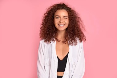 Uçan kıvırcık saçlı güzel kızıl saçlı kız pembe arka planda kameraya bakıp gülümsüyor.