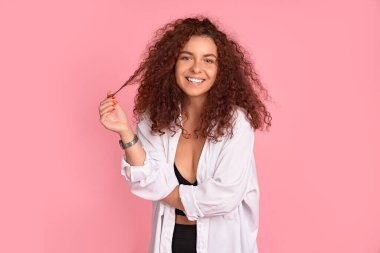 Güzellik, gençlik ve kuaförlük konsepti. Çekici ve neşeli kızıl saçlı, uzun güzel saçları ve doğal rengi olan, kameraya nazikçe gülümseyen, iplikçiklerle oynayan, pembe duvarın üzerinde poz veren bir kadın..