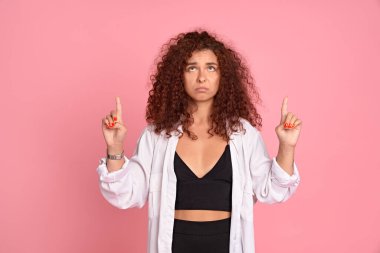 Dost canlısı görünümlü üzgün beyaz saçlı, günlük kıyafetler giyen, işaret parmağı kaldıran, yön gösteren, pembe arka planda reklamını yapan kızıl saçlı kız..