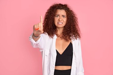 Çekici ve neşeli kızıl saçlı uzun güzel saçları ve doğal rengi olan kadın kamerayı işaret ediyor. Temizlik ve düzen, kameraya tiksindirici bir şekilde, pembe duvarın üzerinde poz vermek..