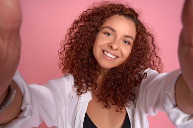 Çekici, mutlu, kıvırcık saçlı, kıvırcık saçlı bir kadın akıllı telefon tutuyormuş gibi elini öne çekiyor, kameraya selfie çekiyor, neşeyle sırıtıyor, turist turistik yerleri geziyor..