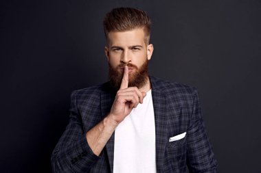 Takım elbiseli yakışıklı sakallı adam sessizlik istiyor. O, parmağını dudaklarına kaldırıyor. Adam dikiliyor ve gizlice kameraya bakıyor. Siyah arkaplanda izole.