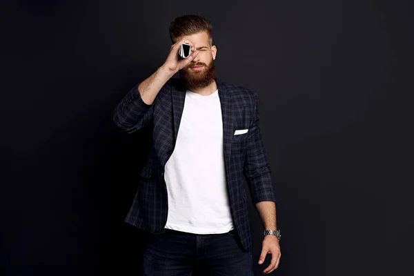 Confident businessman with ginger beard standing on a black background holding a newspaper near his face like a spyglass.