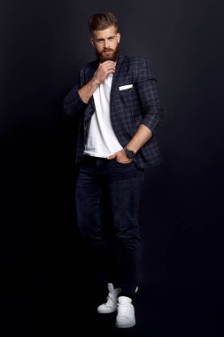 Kendinden emin ve yakışıklı. Zencefilli sakallı, şık giyinmiş, siyah arka planda poz verirken kameraya bakan genç bir adam..