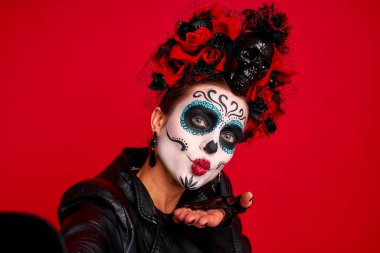 Komik folklor cadısı yaratığın yakın plan fotoğrafı. Karakterin ölüm günü, korkunç makyaj maskeli balosu. Kafatası şekeri kızı selfie çeker. Çiçekli başlık giyer. Deri jaket kırmızı arka planda izole olur..