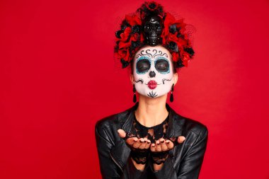 Kafasında çiçek çelengi, kafatasında kafatası ve siyah eldivenleri olan şeker kafalı genç bir kadın kameraya öpücük yolluyor. kırmızı arkaplanda izole edilmiş. Cadılar Bayramı konsepti.