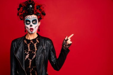 Kafasında çelenk, kafasında kafatası ve siyah eldivenleri olan şeker kafalı genç bir kadın boş bir alanı işaret ediyor. kırmızı arkaplanda izole edilmiş. Cadılar Bayramı veya Calavera Catrina konsepti.
