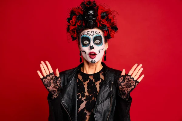 Young woman with a sugar skull makeup with a wreath of flowers on her head and a skull raises her hands up in fear in black gloves. isolated on red background. halloween concept.