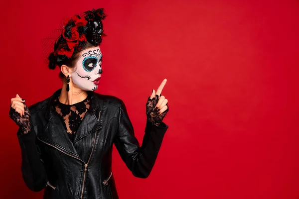 Young woman with sugar skull makeup with a wreath of flowers on her head and skull and black gloves points to a free space. isolated on red background. concept of Halloween or Calavera Catrina.