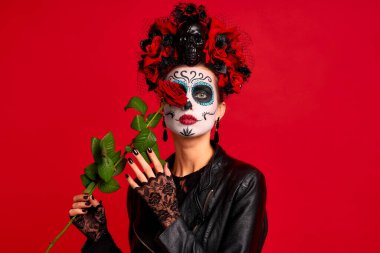 Kafasında ve kafasında çiçek çelengi olan kafatası şekeri makyajlı romantik ölü bir kız kırmızı arka planda kırmızı gül tutan siyah eldivenler giyiyor. Cadılar Bayramı veya Calavera Catrina konsepti.