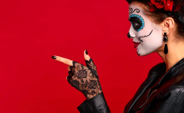Young woman with sugar skull makeup with a wreath of flowers on her head and skull and black gloves points to a free space. isolated on red background. concept of Halloween or Calavera Catrina.