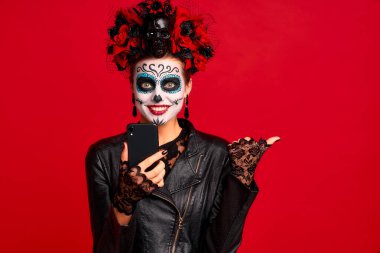 Girl with sugar skull makeup with a wreath of flowers on her head and skull, wearth lace gloves and leather jacket, shocked with news from her phone isolated on red background. clipart