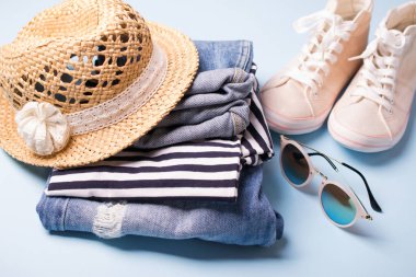 Bayan yazlık giysi ve aksesuarları yığını. Kot tulum, gözlük, spor ayakkabı, hasır şapka, çizgili üst. Seyahat için rahat giysiler