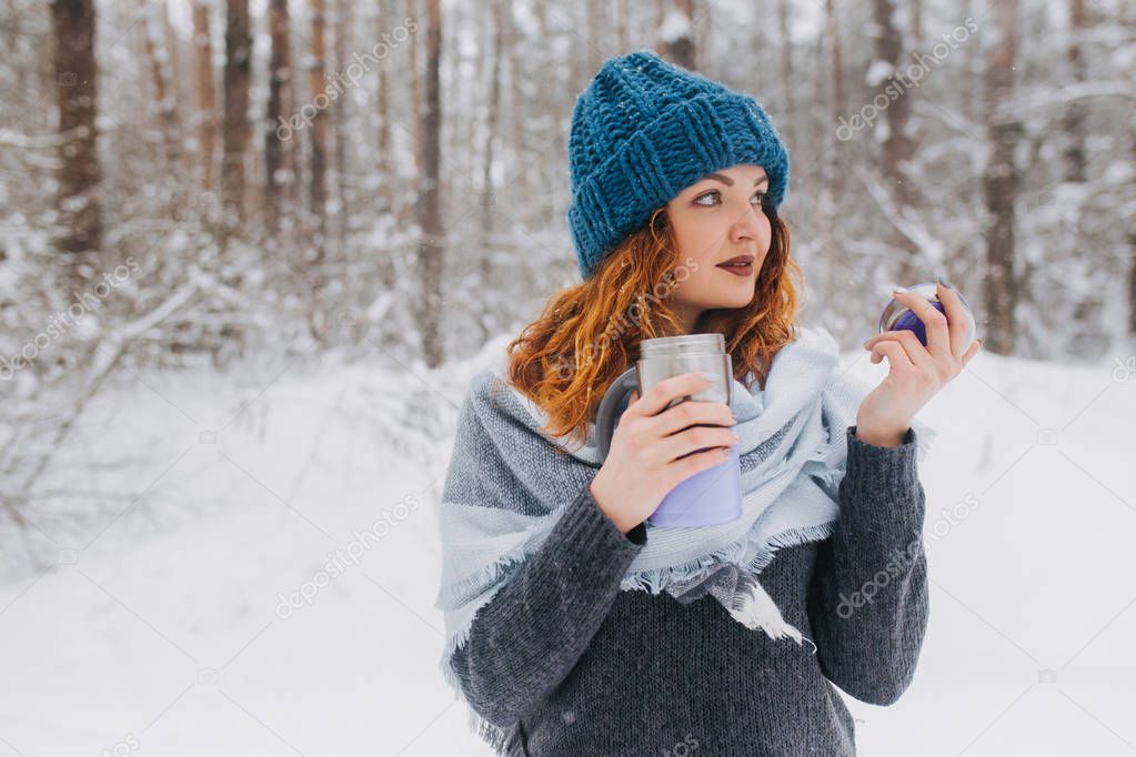 Girl in a hat and a sweater holding cup of tea  in the forest with snow