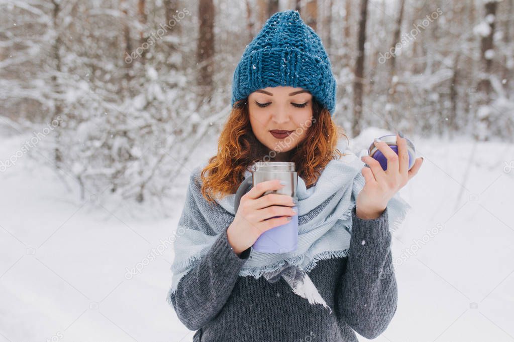 Girl in a hat and a sweater holding thermocup with tea in the forest with snow