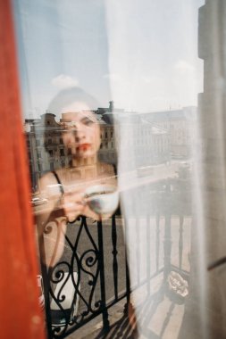 Genç ve güzel bir kadın, bir fincan mis kokulu kahveyle pencerede duruyor.