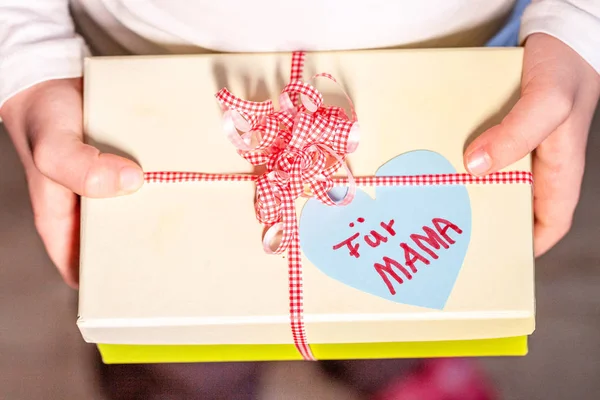 Little child holding gift box with ribbon and heart for her mum - Translation: for mum