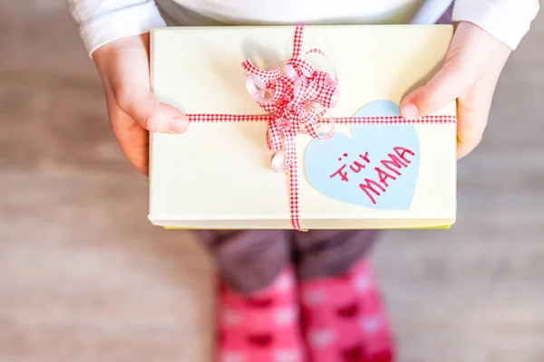 Little child holding gift box with ribbon and heart for her mum - Translation: for mum