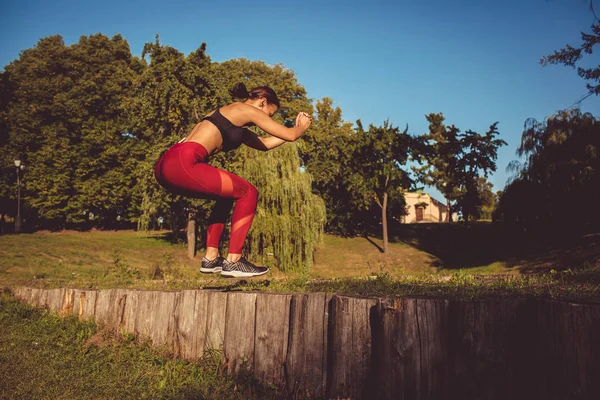 Caucasian woman in red leggings and sportive top warming up in park on wooden poles