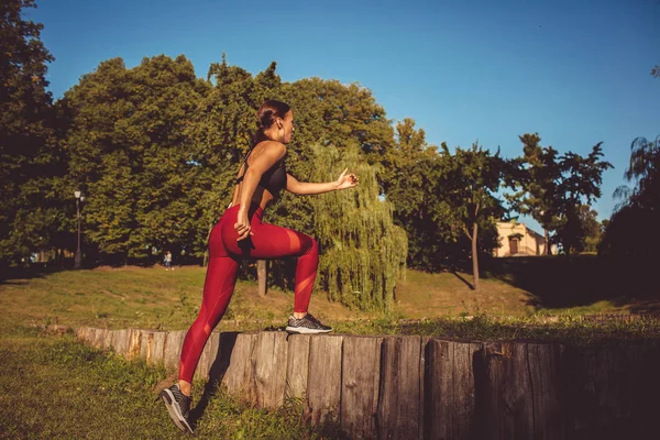 Caucasian woman in red leggings and sportive top warming up in park on wooden poles