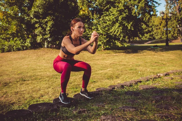 woman in red leggings and sportive top doing squats in park meadow