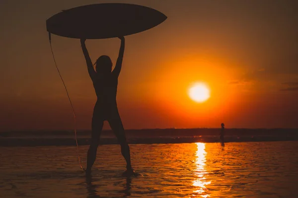 surfer woman with surfboard above head, sea water and sunset in sky
