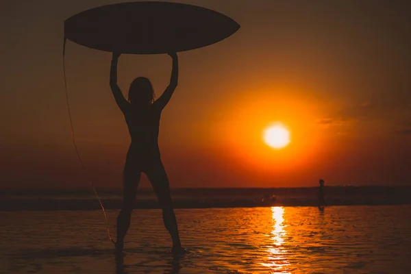surfer woman with surfboard above head, sea water and sunset in sky