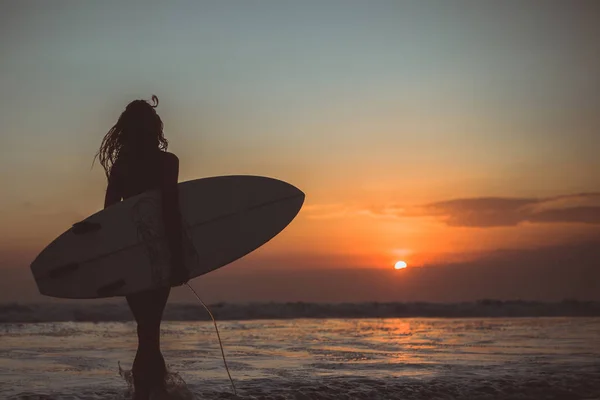 back view of woman silhouette holding surfboard in sea water at sunset
