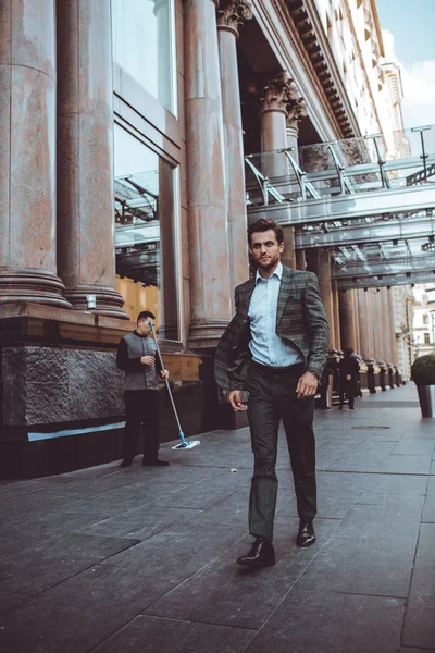 full length of stylish Caucasian gentleman in tweed suit jacket walking in street at building