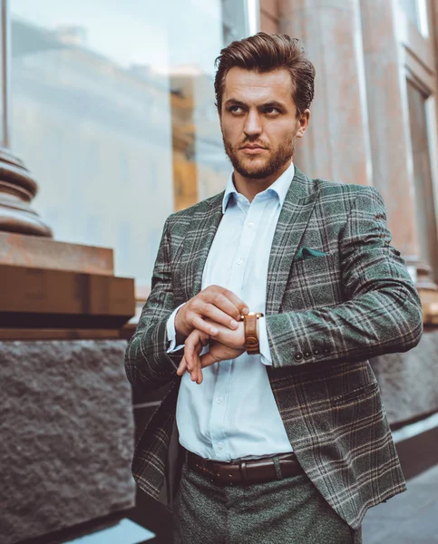 serious elegant Caucasian gentleman in tweed suit jacket walking in street at building and checking time on wrist watch