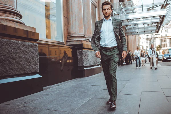 full length of stylish Caucasian gentleman in tweed suit jacket walking in street at hotel building
