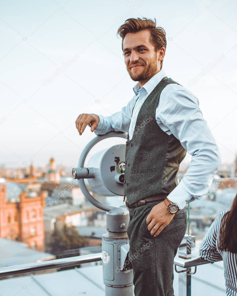 smiling stylish Caucasian man in suit Vest standing at city view telescope on roof 